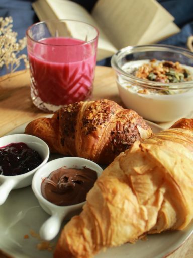 A plate with two croissants, two small bowls of jam, and a glass ft. a red smoothie