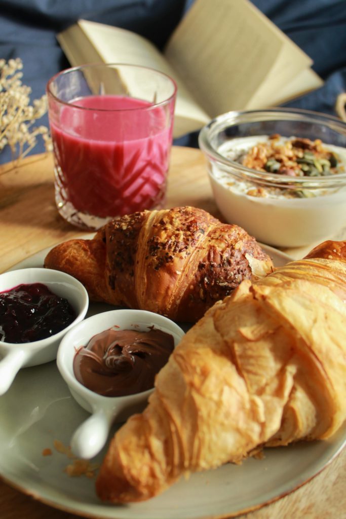 A plate with two croissants, two small bowls of jam, and a glass ft. a red smoothie