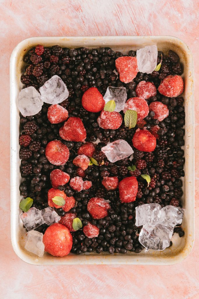Various frozen berries on a sheet pan, with ice cubes and strawberry leaves sprinkled about for decor